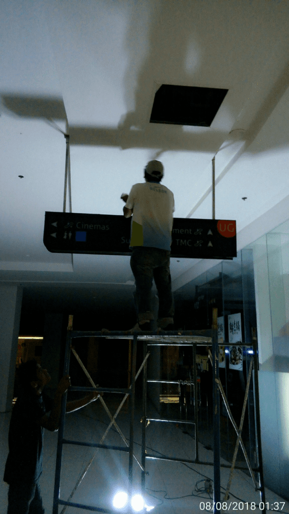 Acrylic signage installation in Makati: worker on scaffolding, assistant below in dim indoor space, by M&G Global Ads.